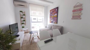 A modern living room with a sofa, a dining table, and a TV. There is a window with blinds, a painting on the wall, and a plant on a shelf.