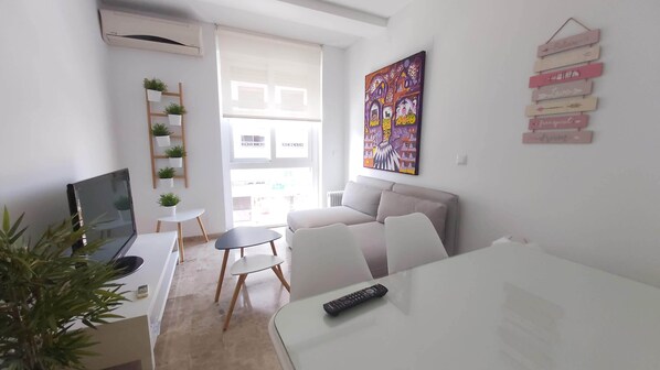 A modern living room with a sofa, a dining table, and a TV. There is a window with blinds, a painting on the wall, and a plant on a shelf.