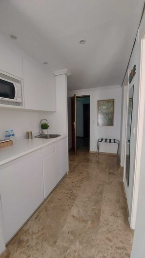 A narrow hallway with a kitchen area on one side, featuring white cabinets, a microwave, and a sink.