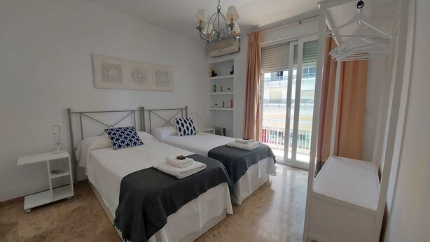 A clean, modern bedroom with two beds, a chandelier, a small table, and a balcony with curtains.