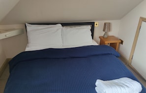 Un letto singolo con cuscini bianchi e coperta blu, un comodino con una lampada e un piccolo tavolo in legno.