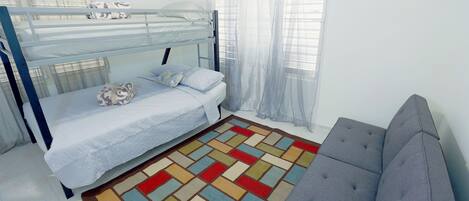 A bunk bed with a patterned blanket, a grey sofa, and a colorful rug in a room with white walls and a window with sheer curtains.