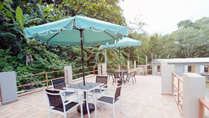 Un patio al aire libre con mesas y sillas, un paraguas verde y vistas de la vegetación circundante.