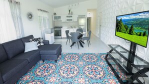 Una sala de estar moderna con un sofá, un tapete estampado, un mueble de televisión de vidrio y una cocina al fondo.