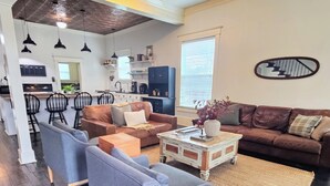A modern living room with a brown leather sofa, a blue armchair, and a coffee table. There is a kitchen in the background with white cabinets and a black countertop. - The Conner House-Walk to Magnolia! (Waco)