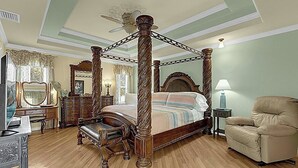 Een hemelbed met houten frame en baldakijn, een chaise longue, een kaptafel met spiegel, en een lamp op een bijzettafeltje.