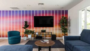 A modern living room with a flat-screen TV mounted on a wall with a gradient sunset design, two blue sofas, a round coffee table, and potted plants. - Revolution Retreat- Heated Pool 5 Mins to Old Town-Hidden Room (Scottsdale)