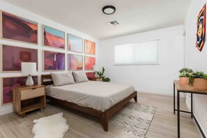 Une chambre à coucher avec un lit en bois, plusieurs œuvres d’art encadrées accrochées au mur, une table de chevet avec une lampe et une petite table avec des plantes.