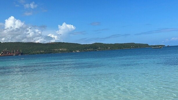 On aperçoit clairement une plage avec une eau peu profonde, une côte lointaine et quelques bateaux.