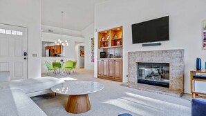 A modern living room with a fireplace, a dining area with green chairs, and a kitchen visible through an open door. - Modern Desert Retreat: Luxurious Comfort & Endless Activities (Palm Desert)