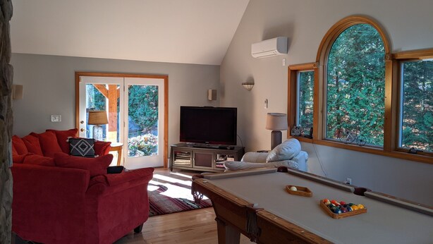 Ein Wohnzimmer mit Billardtisch, einem roten Sofa, einem Fernseher und großen Fenstern mit Blick auf Bäume.