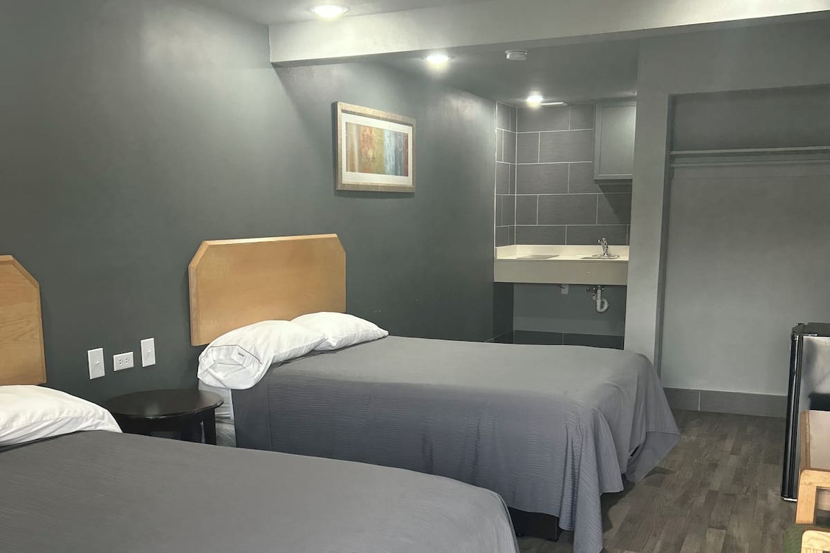 A room with two beds, a small table, a bench, a painting on the wall, and a bathroom with a sink and mirror.