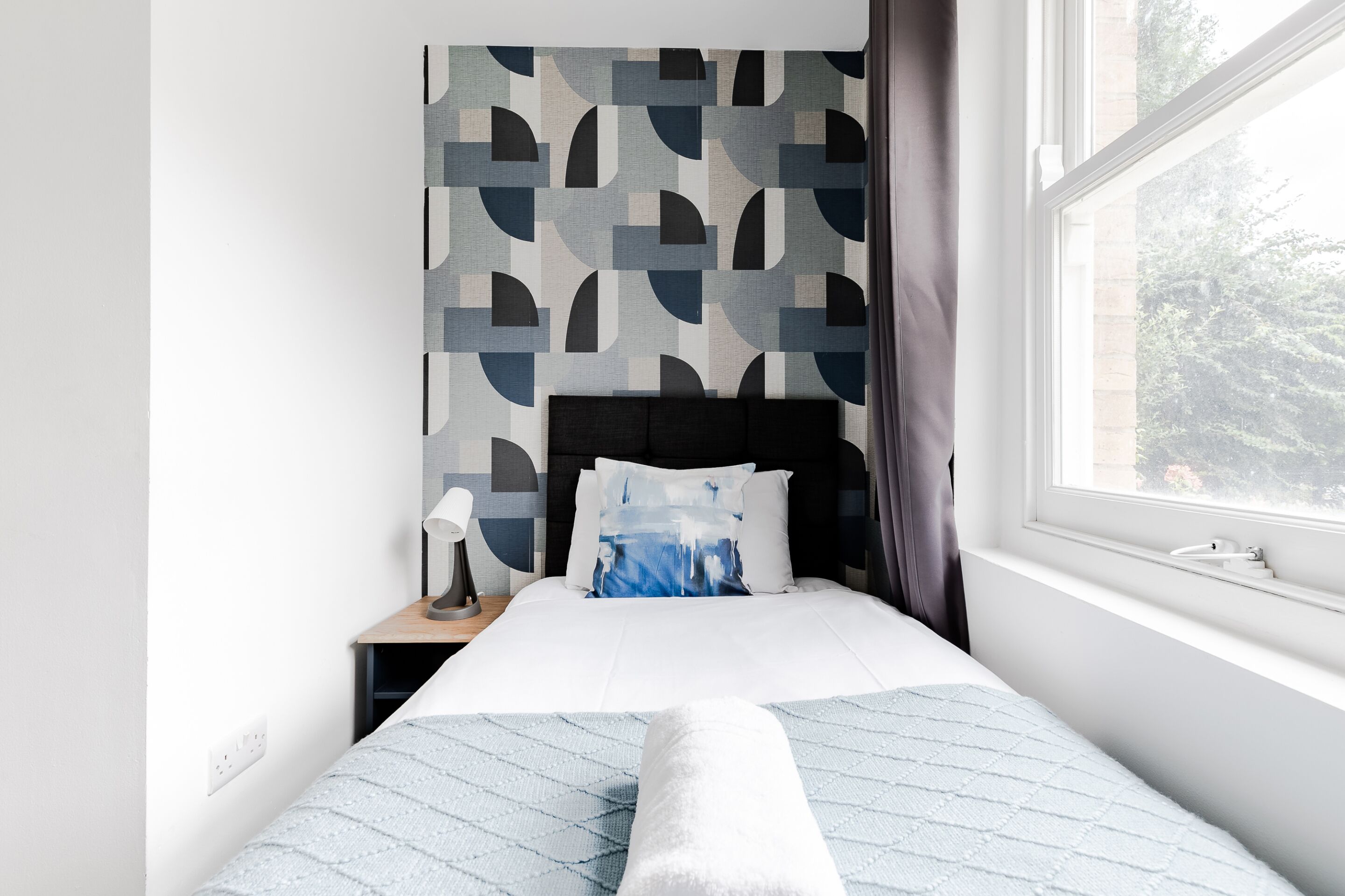 A neatly made bed with a white and blue quilt, a nightstand with a lamp, and a window with a view of trees.