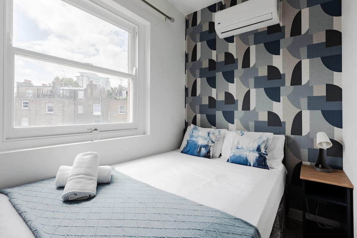 A bedroom with a large window, a bed with blue and white bedding, and a geometric-patterned wall.