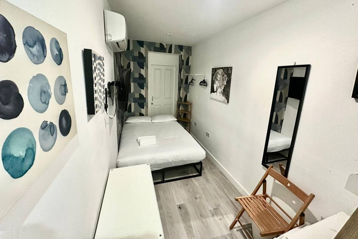 A compact hotel room with a bed, a small desk, a chair, a mirror, and a wall with abstract art.