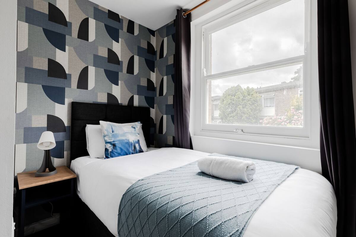 A hotel room with a bed, a nightstand with a lamp, a window with curtains, and a geometric-patterned wall.