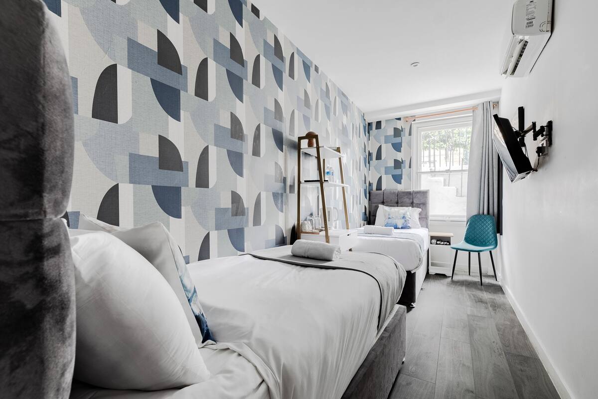 A modern hotel room with a geometric-patterned wall, a bed with white bedding, a small desk with a chair, and a window with curtains.