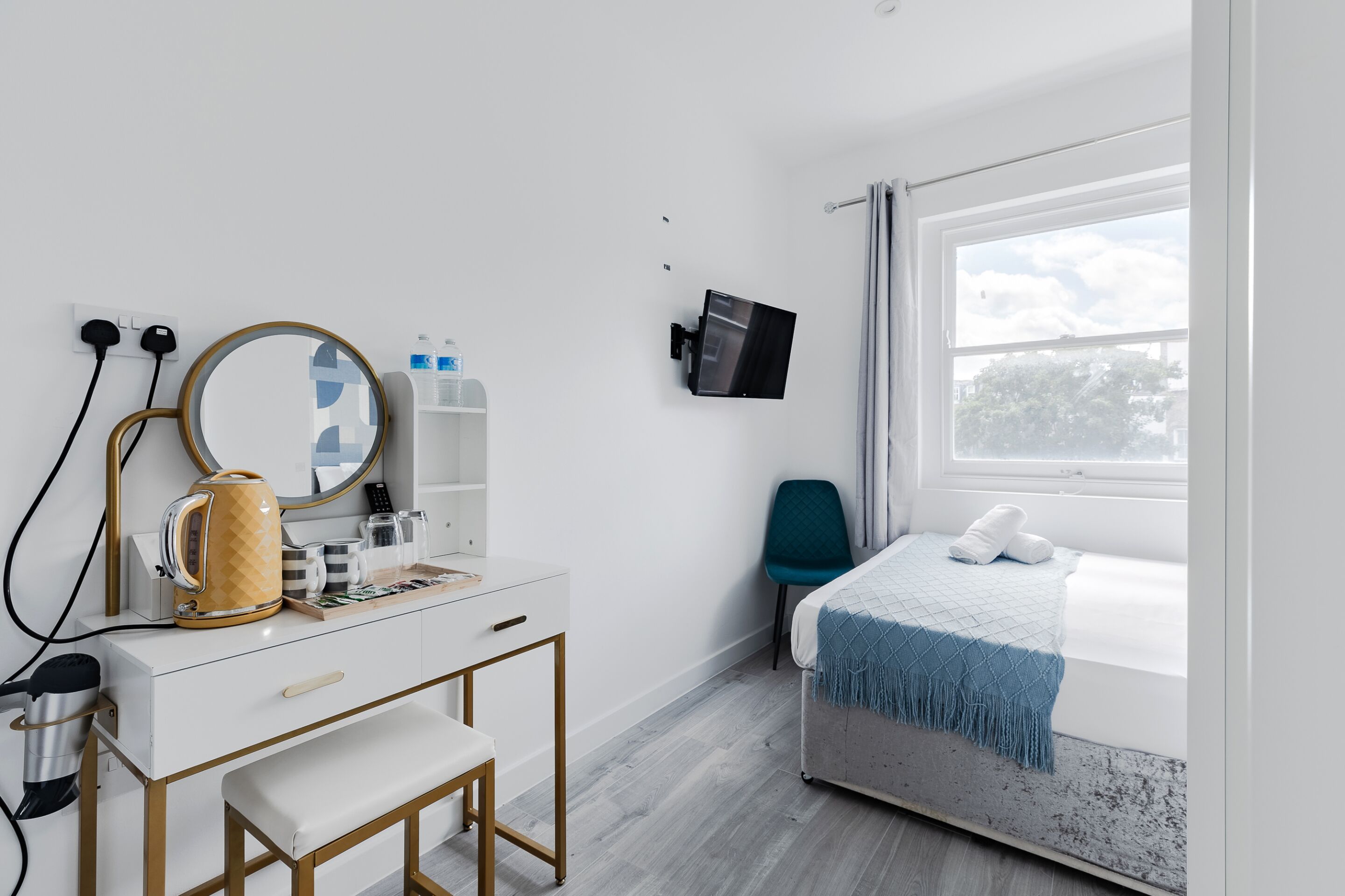 Een moderne hotelkamer met een witte dressoir, een ronde spiegel, een flatscreen-televisie, een bed met een blauwe deken, en een raam met uitzicht op bomen.