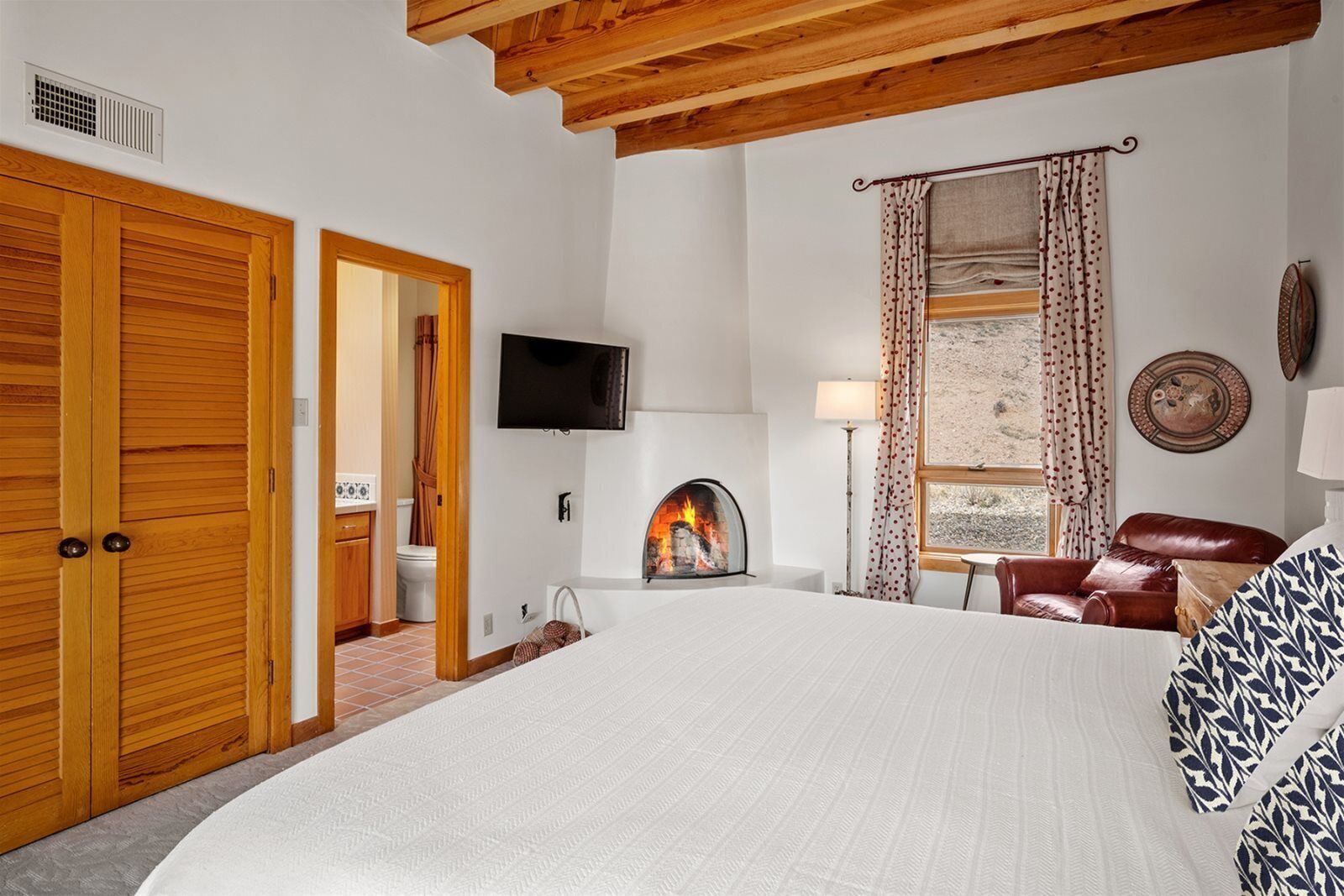 A bedroom with a large bed, wooden wardrobe, a fireplace, a TV, and a window with curtains.