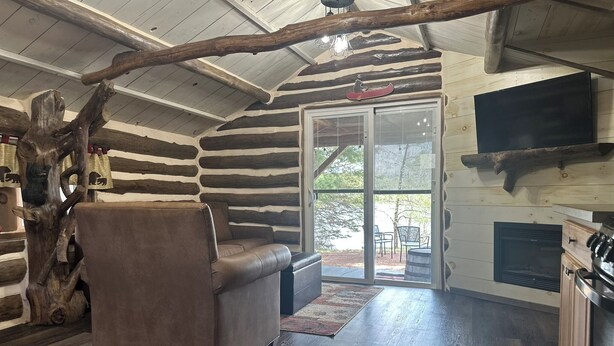 A log cabin interior with a leather sofa, wooden beams, a large tree stump, and a sliding glass door leading outside. - Log Cabin on Lake Alice & ATV/UTV Trails, Papa Bear 2-Bedroom (Tomahawk)