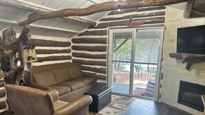A log cabin interior with a leather sofa, a fireplace, and a sliding glass door leading to an outdoor patio. - Log Cabin on Lake Alice & ATV/UTV Trails, Papa Bear 2-Bedroom (Tomahawk)