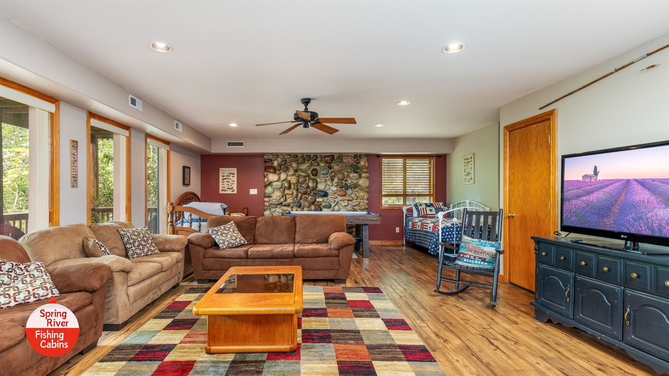 A spacious living room with a large television, comfortable seating, a ceiling fan, and a stone wall.