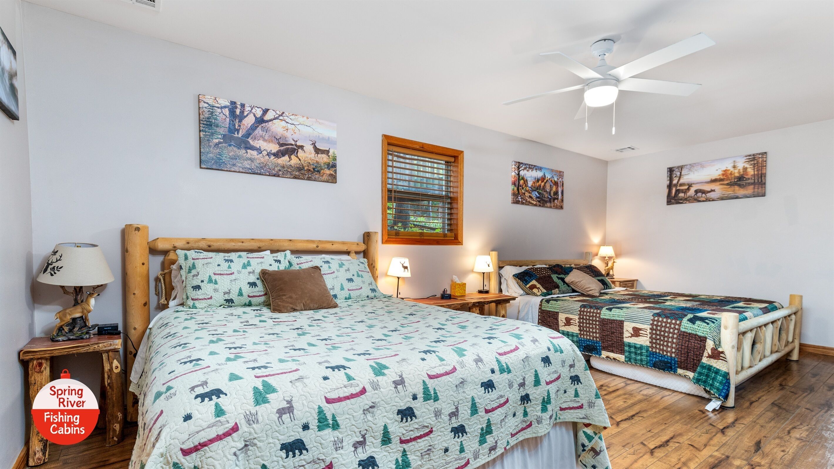 A bedroom with a large bed, a smaller bed, wooden headboards, two nightstands, a ceiling fan, and two wall-mounted paintings.