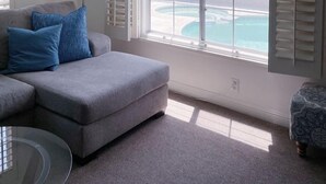Un salon moderne avec un canapé gris, des coussins bleus, une table basse en verre et une vue sur une piscine et des montagnes, le tout visible à travers de grandes fenêtres équipées de volets.