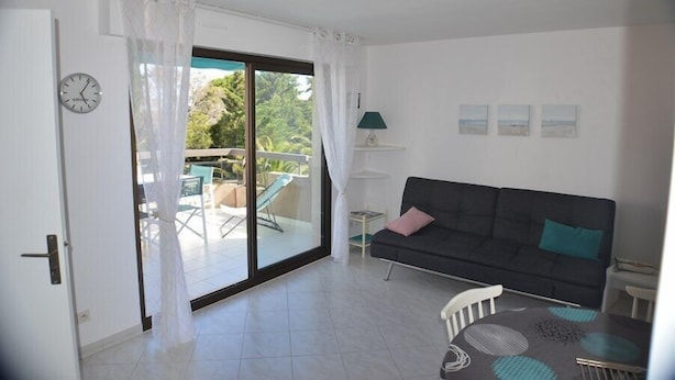 A room with a sliding glass door leading to an outdoor area with chairs and a table. There is a dark-colored sofa with cushions, a small table with a lamp, and a clock on the wall.