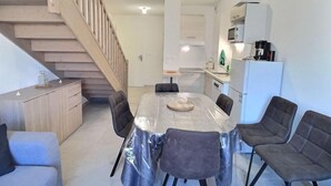 A compact living space with a dining table covered in plastic, surrounded by chairs, a staircase, a kitchen area with a refrigerator, and a microwave. - Appartement Confortable Près de la Plage (Sérignan)
