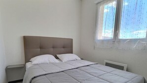 A bedroom with a bed, a window with curtains, and a radiator. - Appartement Confortable Près de la Plage (Sérignan)