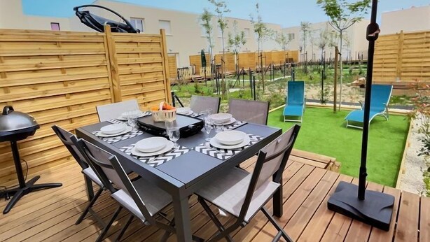 Un patio con una mesa dispuesta para cuatro personas, rodeado de piso de madera, un paraguas rojo y dos sillones azules.