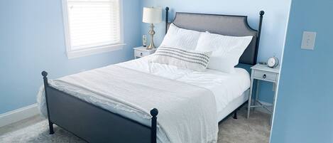 A neatly made bed with white linens and a patterned throw, a bedside table with a lamp and a clock, and a window with blinds.