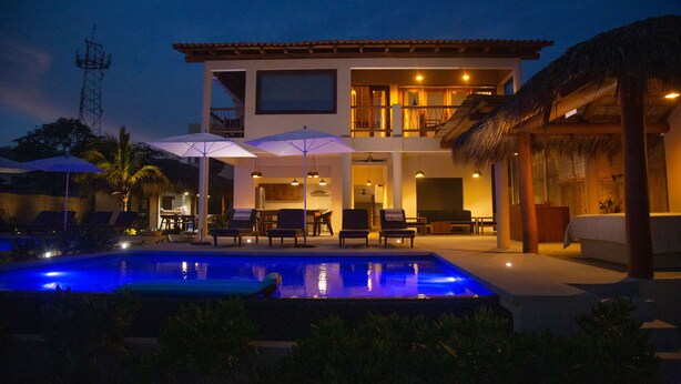 Un hotel de dos pisos con piscina, mobiliario exterior y una estructura con techo de paja.