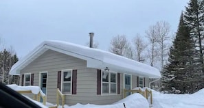 Une petite maison à un étage, avec un toit en pente recouvert de neige, entourée d’arbres.