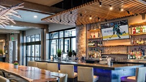A modern bar area with a wooden bar counter, bar stools, and a large screen displaying a hockey game.