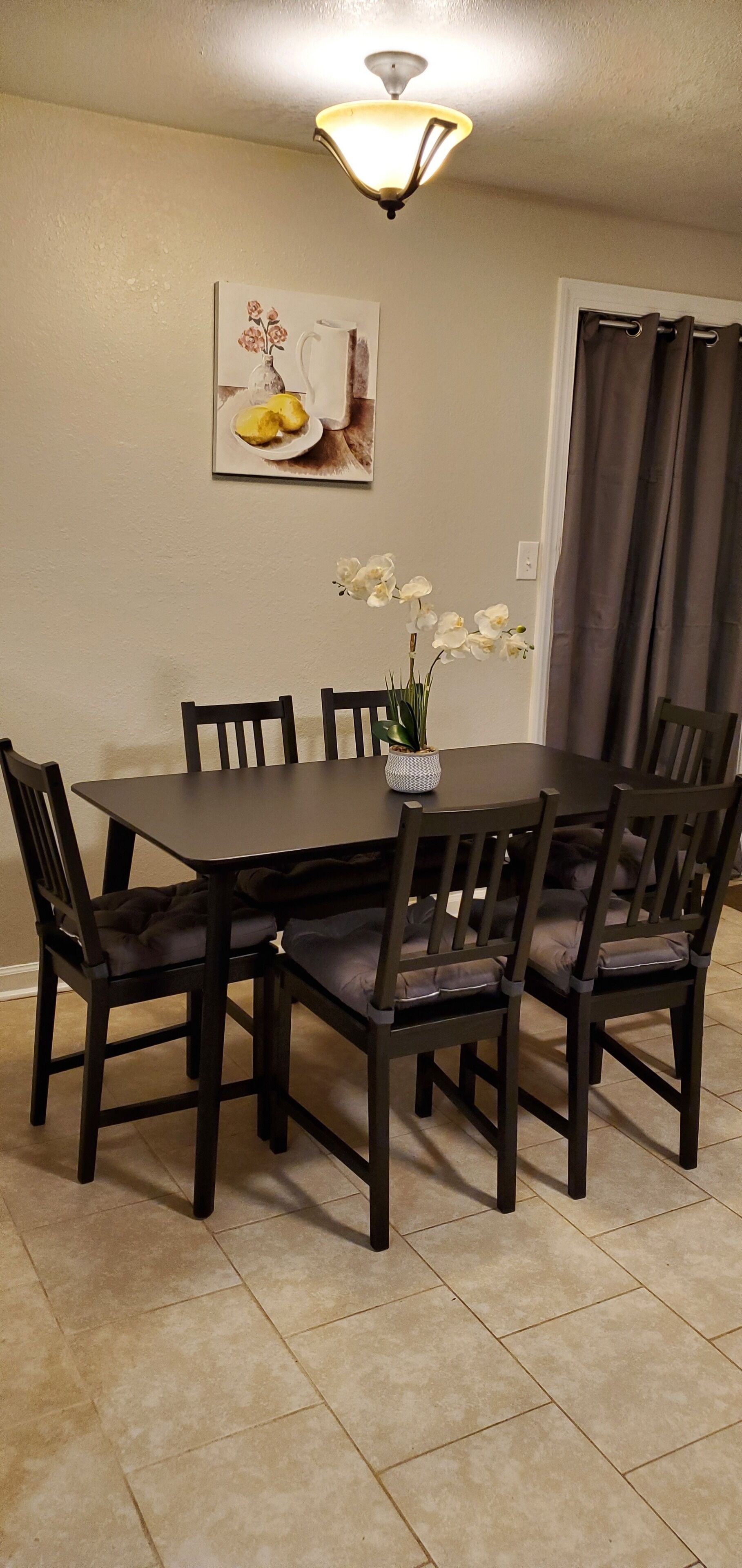 A dining area with a dark wooden table and chairs, a vase with white flowers, and a wall-mounted picture.