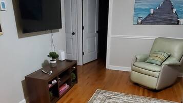 A living room with a flat-screen TV, a wooden entertainment unit, a green armchair, and a patterned rug.