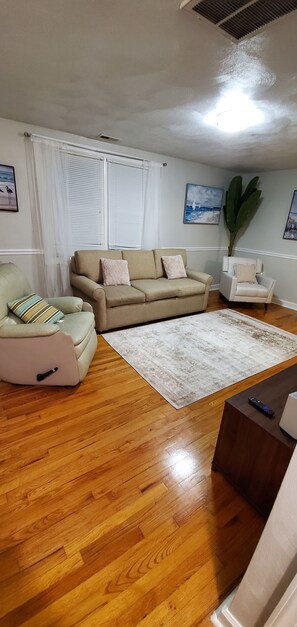 Una sala de estar con un sofá color beige, un sillón verde y un sillón blanco. Se observa una planta grande, piso de madera y una rejilla en el techo.