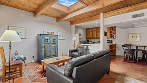 A spacious living area with a high wooden ceiling, a skylight, a sofa, a coffee table, a lamp, and a blue cabinet. - Fort Marcy 90 | Walk to Plaza | Full Kitchen | Private Bedroom | Hot Tub | Pool | Single Level (Santa Fe)