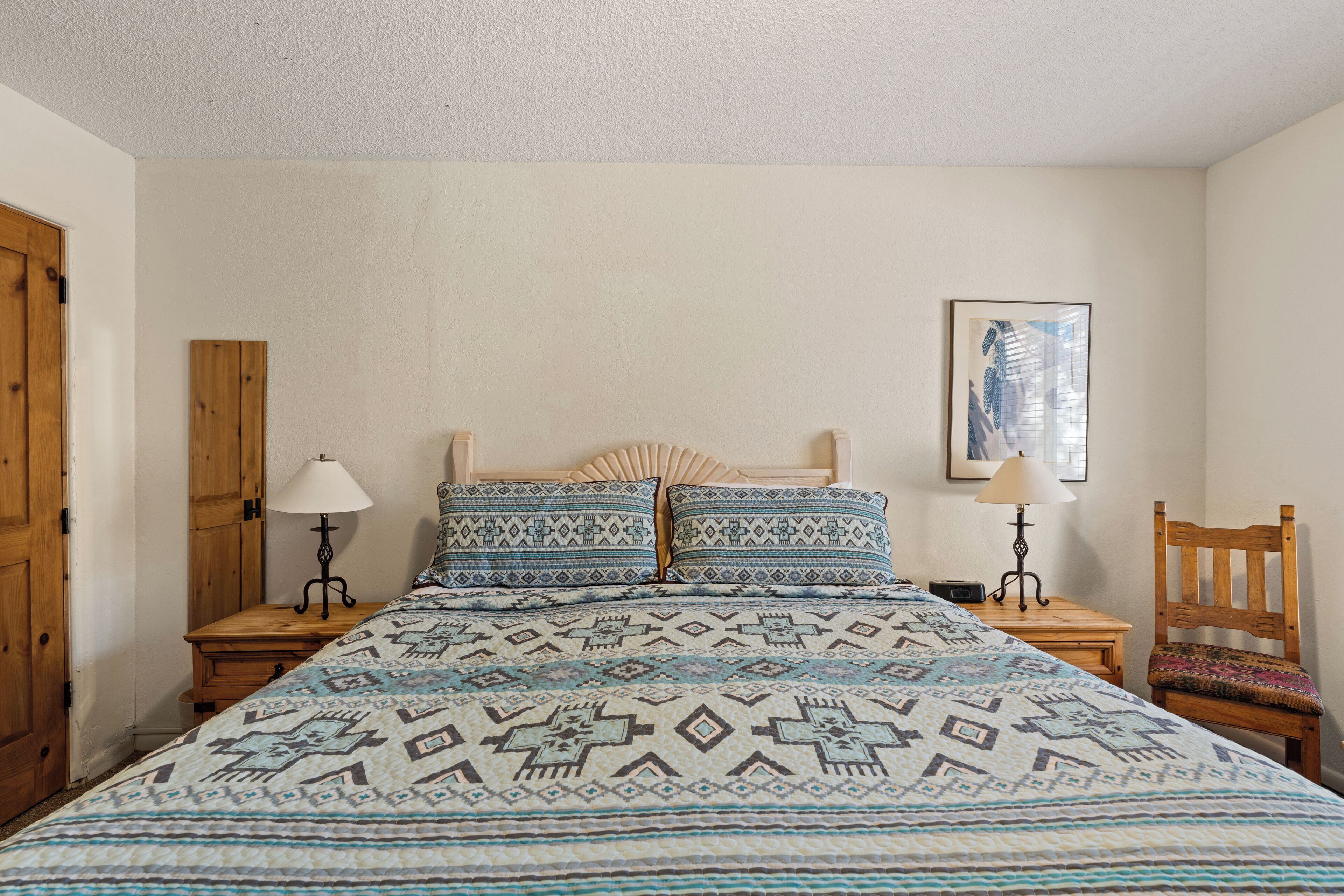 A bedroom with a bed, two nightstands, a wooden chair, a wall-mounted lamp, and a framed picture.