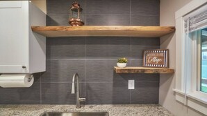 A kitchen with a stainless steel sink, dark tiled backsplash, wooden shelves, and a welcome sign. - Riverfront Cottage with Hot Tub 1/4 mi from Beach (Brookings)