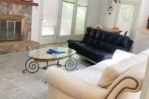 Un salon avec une cheminée en pierre, un canapé en cuir noir, un fauteuil blanc et une table basse en verre.