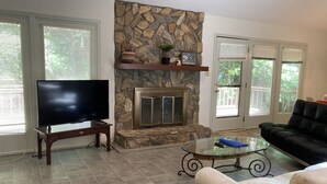 Una sala de estar con chimenea de piedra, un televisor de pantalla plana sobre un mueble bajo, una mesa de centro de vidrio y un sofá negro.