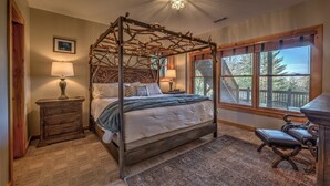A four-poster bed with a wooden frame, a nightstand with a lamp, a bench, and a view of a deck through large windows.
