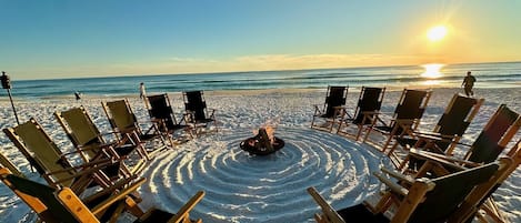 A beach with arranged lounge chairs and a fire pit in the center, overlooking the ocean.