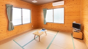 Una habitación con paredes de madera, dos ventanas grandes con cortinas, una mesita con una bandeja, un televisor sobre un mueble y esteras de tatami en el suelo.