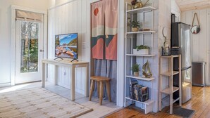 A room with a wooden floor, a white door, a table with a painting, a stool, a shelf with books and plants, and a refrigerator. - Romantic Forest Cabin - private, near everything (Bar Harbor)