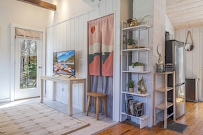 Une pièce avec un plancher en bois, une porte blanche, une table sur laquelle est posée une peinture, un tabouret, une étagère avec des livres et des plantes, et un réfrigérateur.