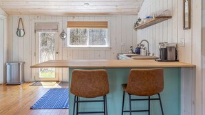 A kitchen with a wooden ceiling, a window with blinds, a sink, a coffee maker, and two bar stools. - Romantic Forest Cabin - private, near everything (Bar Harbor)